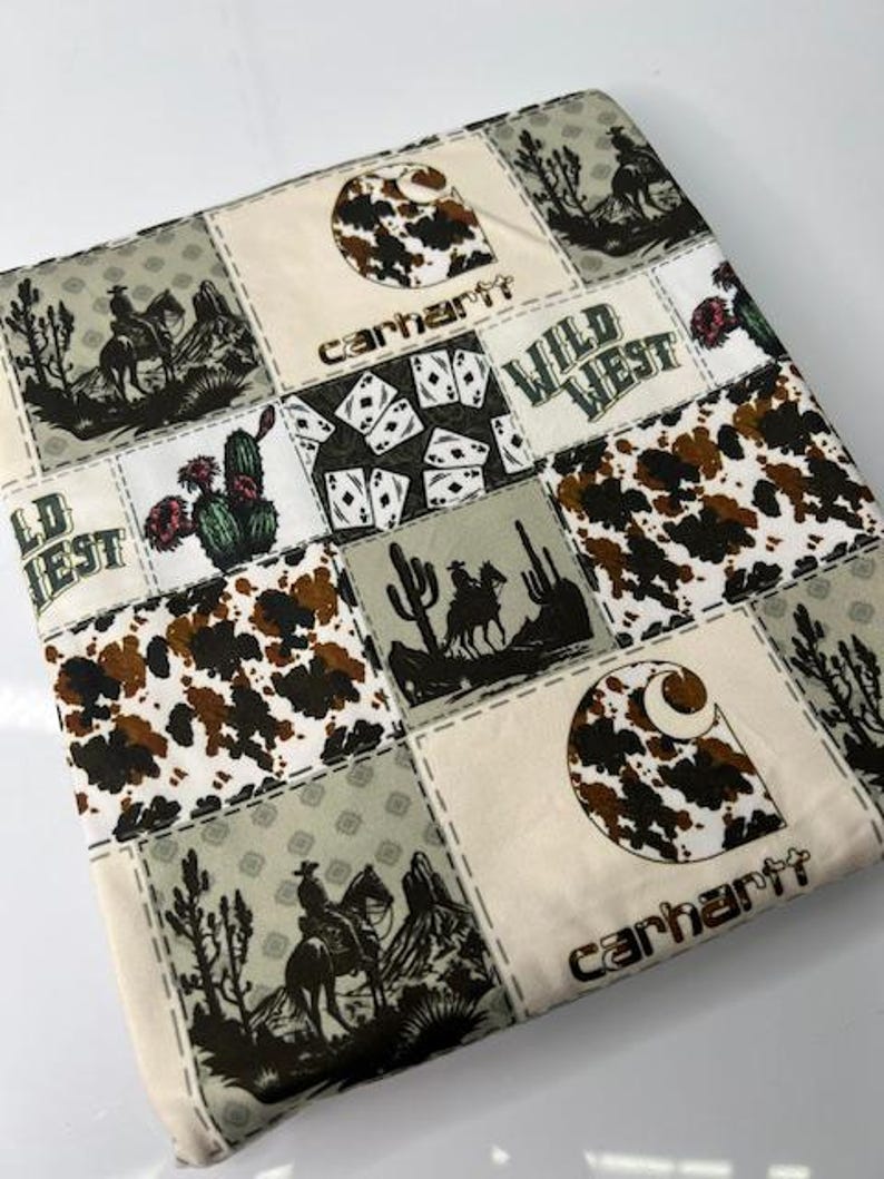 Op de afbeelding: Een patchwork stof met een witte achtergrond en verschillende patronen, waaronder een bruin en wit koeienprint, speelkaarten, cactussen en cowboys die op paarden rijden. De stof heeft ook het Carhartt-logo en de woorden "Wild West".