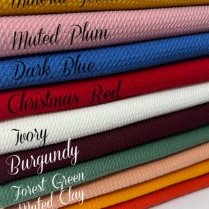May include: A stack of textured fabric swatches in various colors, including yellow, pink, blue, red, white, burgundy, green, peach, orange. Each swatch is labeled with its color name in cursive script.