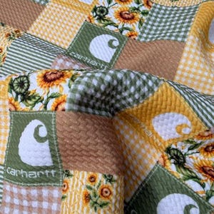 May include: A patchwork quilt featuring squares of various patterns and textures. The quilt incorporates floral prints, gingham, and solid fabrics in shades of yellow, green, and brown. Several squares display the Carhartt logo.