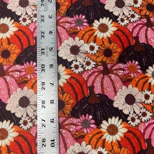 May include: A fabric with a repeating pattern of pumpkins and daisies in shades of orange, pink, and white. The pumpkins have dark brown stems. A ruler is placed vertically along the left side of the fabric, showing measurements in inches.