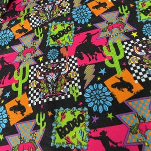 May include: A fabric with a vibrant rodeo-themed pattern. The design features cacti, cowboys, bucking broncos, and the word "Rodeo" in a neon green square. The background is black with pink, orange, blue, and checkered elements.