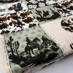 Op de afbeelding: Een witte stof met een patchwork-ontwerp met cowboys, paarden, cactussen en speelkaarten. De stof heeft een bruin, groen en zwart kleurenschema. De tekst "Wild West" en "Carhartt" is zichtbaar op de stof.