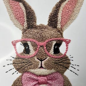 May include: Embroidered brown bunny with pink ears, pink glasses, and a pink bow tie. The bunny is on a white background.