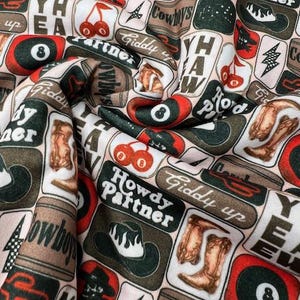 May include: Fabric with a repeating pattern of cowboy-themed illustrations. The design includes cowboy hats, boots, cherries, lightning bolts, and the text "Howdy Partner" and "Cowboys". The color palette features shades of brown, red, black, and white.
