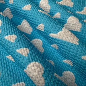 May include: Close-up of a textured, bright blue fabric with a pattern of white clouds. The clouds are irregularly shaped and scattered across the surface. The fabric has a woven appearance, creating a subtle texture.