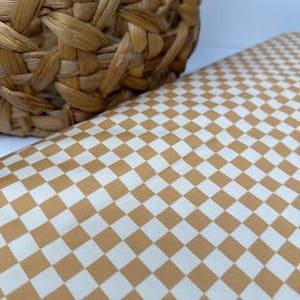 May include: A woven basket made of natural fibers sits next to a fabric with a checkered pattern. The fabric features a repeating design of alternating squares in white and a warm, golden-brown color.