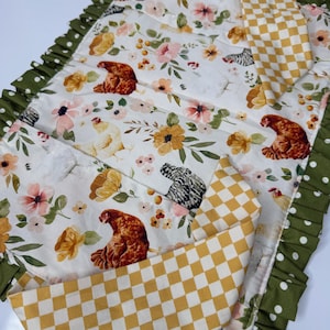 May include: A quilted placemat with a floral and chicken pattern. The placemat features a white background with pink, yellow, and green floral designs, and brown chicken illustrations. A yellow and white checkered pattern is also visible.