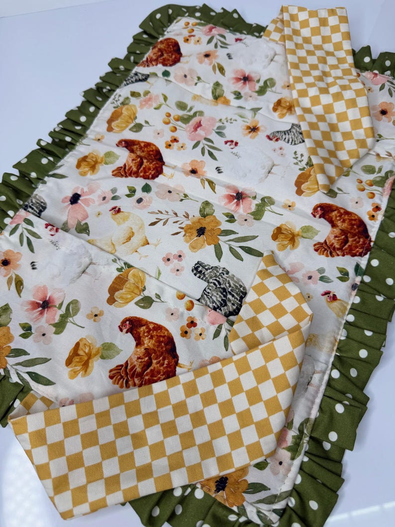 May include: A quilted placemat with a floral and chicken pattern. The placemat features a white background with pink, yellow, and green floral designs, and brown chicken illustrations. A yellow and white checkered pattern is also visible.