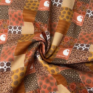 May include: Patchwork fabric with a variety of patterns in shades of brown, orange, and yellow. The design includes floral, leopard print, and gingham squares, with the Carhartt logo on some patches. The fabric is folded, showing its texture.