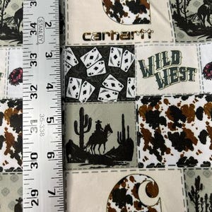 Op de afbeelding: Een veelkleurige stof met een patchwork-ontwerp met een Carhartt-logo, een Wild West-thema en een koehuidpatroon. De stof is beige met bruine, zwarte en witte accenten.