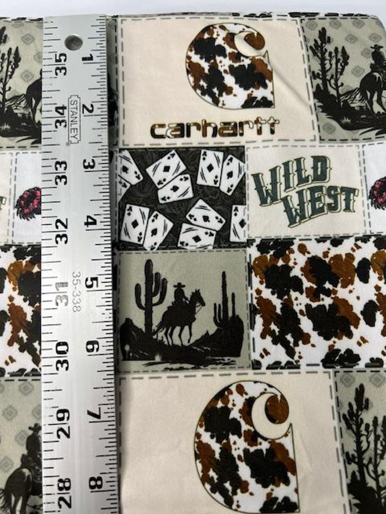 Op de afbeelding: Een veelkleurige stof met een patchwork-ontwerp met een Carhartt-logo, een Wild West-thema en een koehuidpatroon. De stof is beige met bruine, zwarte en witte accenten.