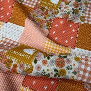 May include: Close-up of a patchwork fabric with a variety of patterns and colors. The design includes floral prints, gingham, and solid squares in shades of orange, peach, and white. The Carhartt logo is visible on a few of the patches.