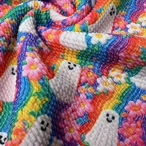 May include: A vibrant fabric with a repeating pattern of smiling white ghosts and colorful flowers. The background features a rainbow of colors, including red, orange, yellow, green, blue, and purple. The fabric has a textured, slightly raised surface.