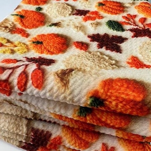 May include: A stack of cream-colored towels with an embroidered autumn design. The towels feature orange pumpkins, brown and orange leaves, and yellow accents. The towels are folded and stacked, showcasing the repeating pattern.