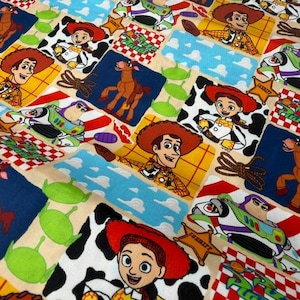 May include: A colorful fabric featuring a patchwork design with various cartoon characters. The design includes images of Woody, Buzz Lightyear, Jessie, and other characters from the Toy Story franchise. The fabric has a mix of colors, including blue, red, yellow, and white.