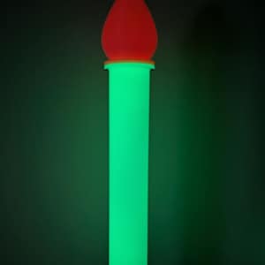 Puede incluir: Una lámpara brillante con forma de vela cilíndrica. La base es de un verde brillante, coronada por una luz roja en forma de llama. El fondo es de color verde oscuro.