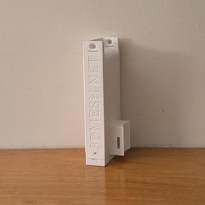 May include: A white 3D-printed object with the text ".3DMESH.NET" on the side. The object has a rectangular shape with a small, attached cuboid. It is placed on a wooden surface against a plain wall.