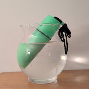 May include: A green cylindrical object with a black cord is partially submerged in water inside a clear, round glass vase. The vase sits on a wooden surface. The object appears to be waterproof.