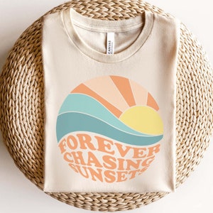 May include: A beige t-shirt with a graphic of a sunset and waves in shades of blue, orange, and yellow. The text "FOREVER CHASING SUNSET" is written in a retro font.