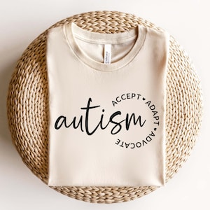 May include: A beige t-shirt with the word "autism" in black lettering. The word is surrounded by a circle with the words "accept", "adapt", "advocate" written around it.
