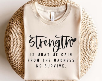 May include: A beige t-shirt with the text "Strength is what we gain from the madness we survive." in black lettering with a small black heart.