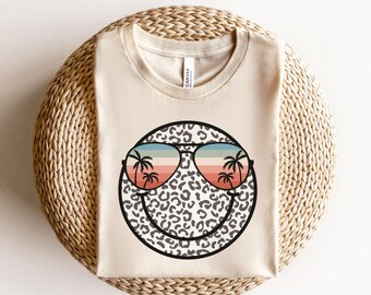 May include: A beige t-shirt featuring a smiling face graphic. The face is printed with a leopard print pattern and has sunglasses with palm trees reflected in the lenses. The sunglasses have a rainbow gradient.