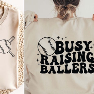 May include: A beige sweatshirt with a black baseball graphic and the text "Busy Raising Ballers" in a retro font.