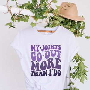 Puede incluir: Camiseta blanca con un gr&aacute;fico morado y blanco que dice "Mis articulaciones salen m&aacute;s que yo."