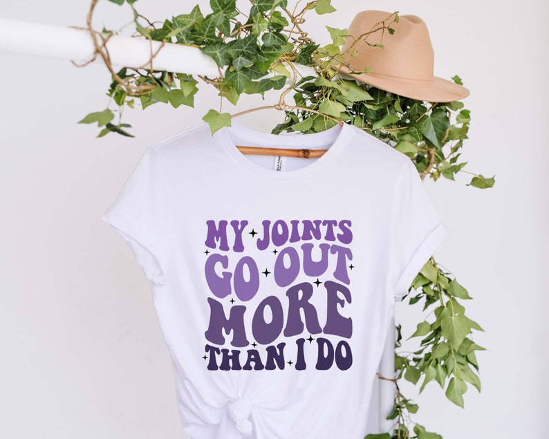Puede incluir: Camiseta blanca con un gr&aacute;fico morado y blanco que dice "Mis articulaciones salen m&aacute;s que yo."