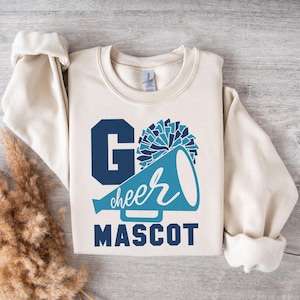 Beige sweatshirt with a cheer-themed design: 'Go Cheer Mascot' in bold blue and white text, a megaphone with 'cheer' in cursive, and a pom-pom graphic. Styled flat on a wooden surface with pampas grass decor for a cozy, aesthetic look