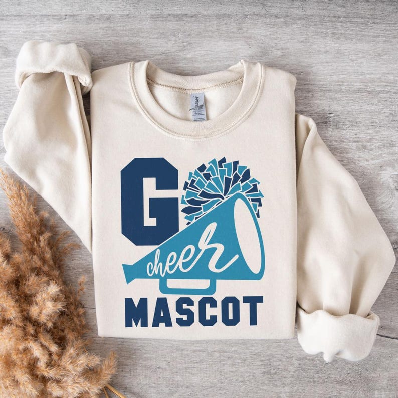 Beige sweatshirt with a cheer-themed design: 'Go Cheer Mascot' in bold blue and white text, a megaphone with 'cheer' in cursive, and a pom-pom graphic. Styled flat on a wooden surface with pampas grass decor for a cozy, aesthetic look