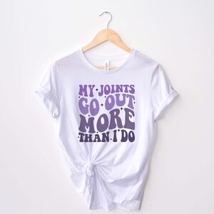 Puede incluir: Camiseta blanca con un gr&aacute;fico morado y blanco que dice "My Joints Go Out More Than I Do" en una fuente retro.