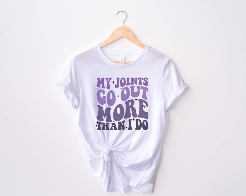 Puede incluir: Camiseta blanca con un gr&aacute;fico morado y blanco que dice "My Joints Go Out More Than I Do" en una fuente retro.