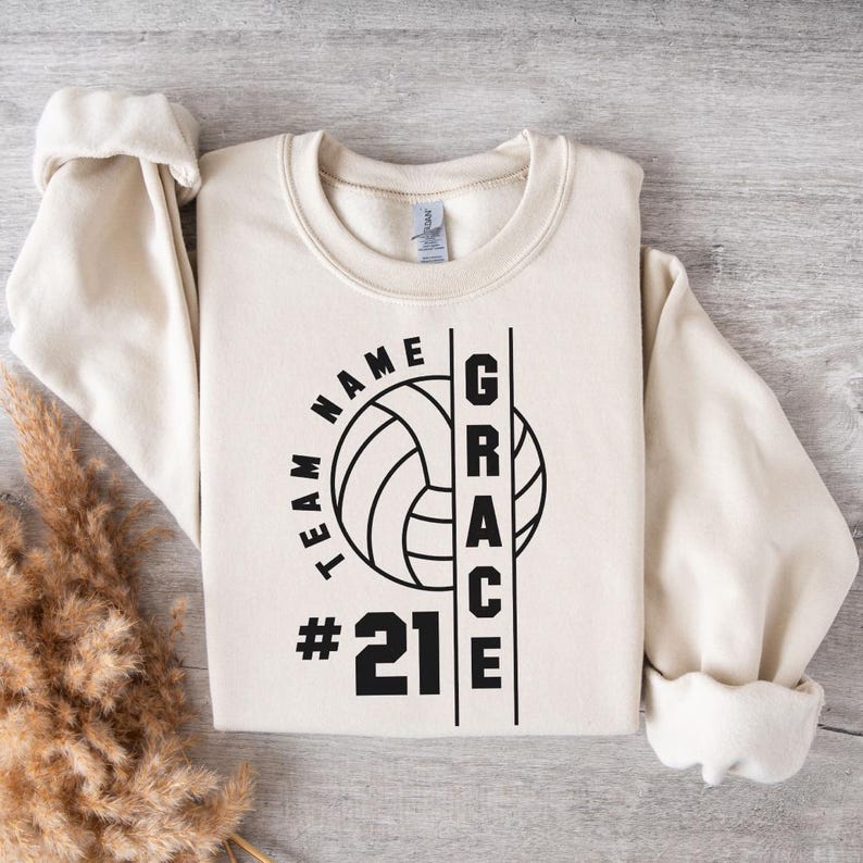 A beige sweatshirt with a custom volleyball design featuring "Team Name" curved around a volleyball icon, "Grace" vertically aligned, and the player number "#21" displayed prominently.