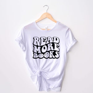 May include: White t-shirt with black text that reads "READ MORE BOOKS" in a retro font with smiley faces in the "O"s of "BOOKS".