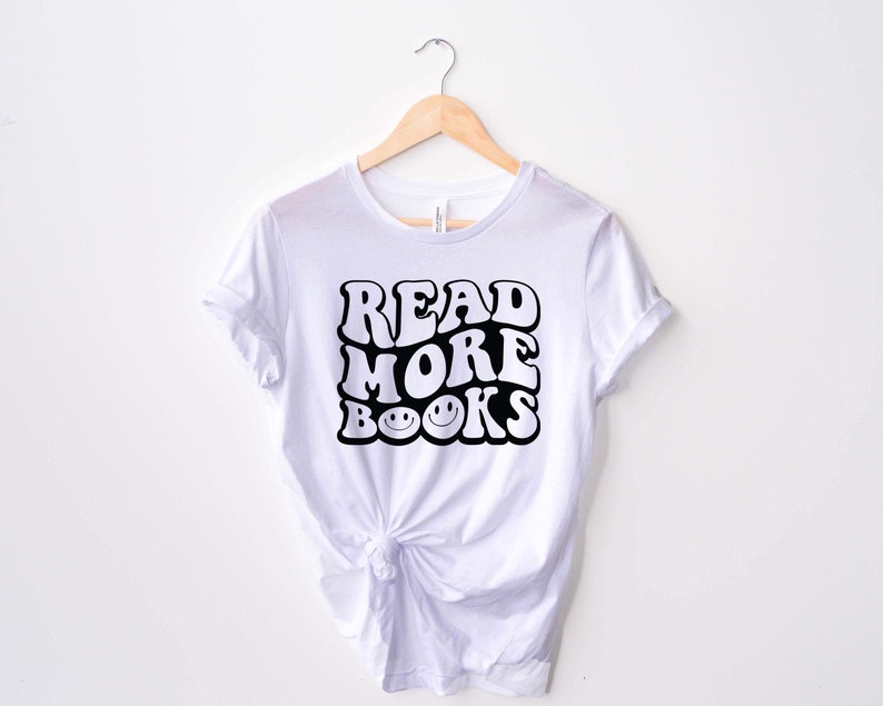 May include: White t-shirt with black text that reads "READ MORE BOOKS" in a retro font with smiley faces in the "O"s of "BOOKS".
