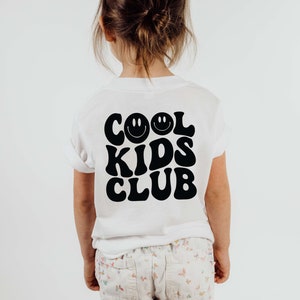 以下が含まれることがあります： 「COOL KIDS CLUB」というレトロなフォントで書かれた黒いグラフィックが施された白いTシャツ。スマイリーフェイスが2つある。