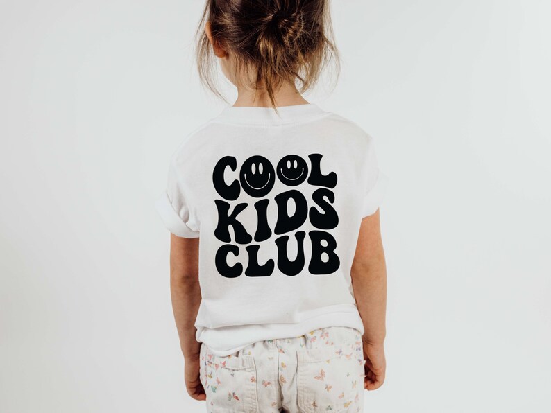 以下が含まれることがあります： 「COOL KIDS CLUB」というレトロなフォントで書かれた黒いグラフィックが施された白いTシャツ。スマイリーフェイスが2つある。