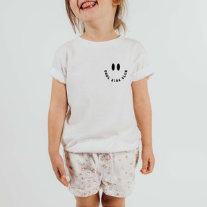 以下が含まれることがあります： 黒いスマイリーと「クールキッズクラブ」の文字が書かれた白いTシャツ。子供がTシャツを着て笑顔を見せています。