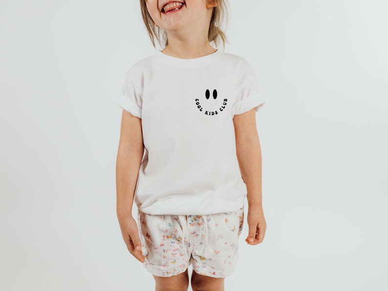 以下が含まれることがあります： 黒いスマイリーと「クールキッズクラブ」の文字が書かれた白いTシャツ。子供がTシャツを着て笑顔を見せています。