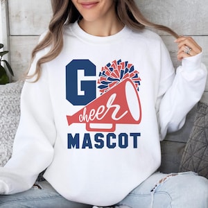 White sweatshirt with 'Go Cheer Mascot' text, featuring a megaphone and pom-pom graphic in blue and red. Worn by a woman in a cozy setting