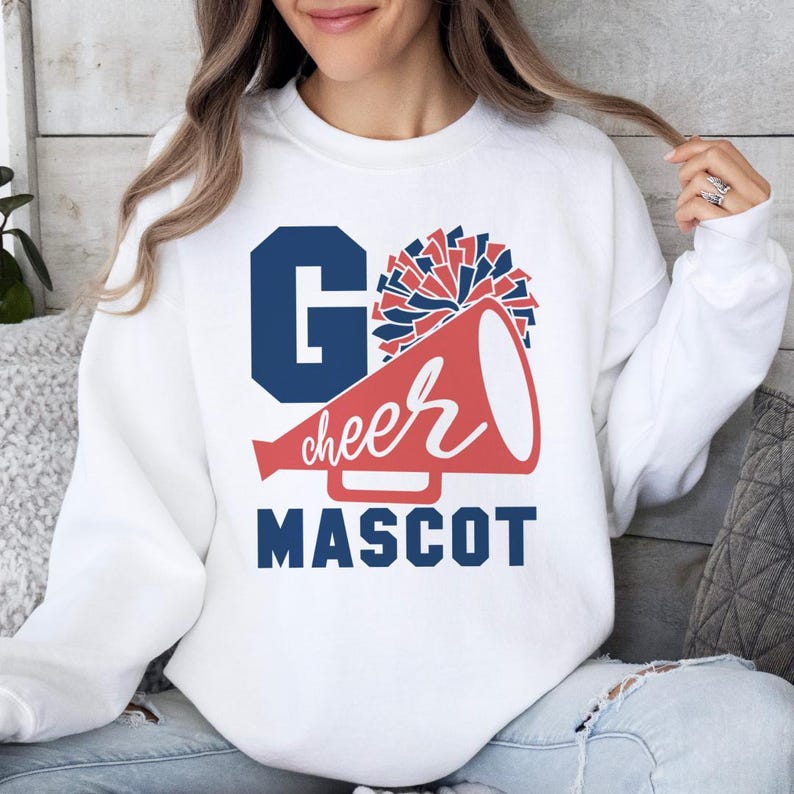 White sweatshirt with 'Go Cheer Mascot' text, featuring a megaphone and pom-pom graphic in blue and red. Worn by a woman in a cozy setting