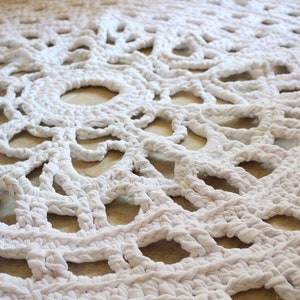 May include: A white, circular, crocheted rug with a detailed openwork design. The rug features a repeating pattern of concentric circles and geometric shapes, creating a textured surface. The rug is made of a thick, soft material.