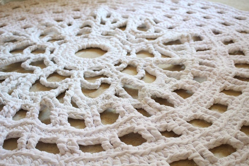 May include: A white, circular, crocheted rug with a detailed openwork design. The rug features a repeating pattern of concentric circles and geometric shapes, creating a textured surface. The rug is made of a thick, soft material.