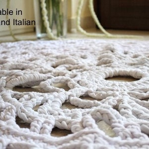May include: Close-up of a white, hand-knitted rug with a textured, openwork design. The rug is made of thick, soft material and features large, circular openings throughout the pattern. The rug is a neutral color and would complement any decor.