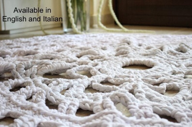 May include: Close-up of a white, hand-knitted rug with a textured, openwork design. The rug is made of thick, soft material and features large, circular openings throughout the pattern. The rug is a neutral color and would complement any decor.