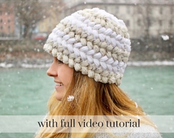 May include: A white and beige crocheted beanie hat with a textured stitch pattern. The hat is worn by a person with long blonde hair, standing in front of a body of water with snow falling.