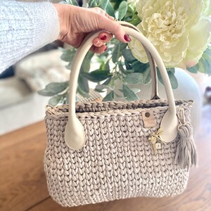 以下が含まれることがあります： 白い革のハンドルと金のチャームが付いたベージュのクロシェ編みのハンドバッグ。バッグは質感のある織り模様です。
