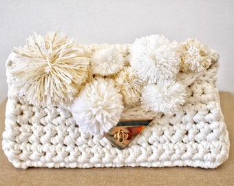 May include: Cream-colored, crocheted clutch with a rectangular shape. The bag is adorned with several fluffy, off-white pom-poms. A gold-colored metal clasp secures the flap. The bag is made of thick, textured yarn, giving it a handmade appearance.