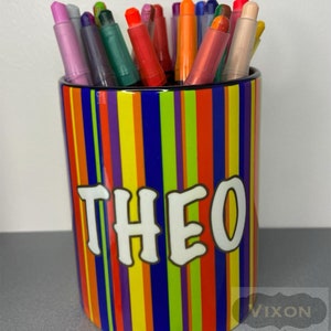 May include: A colourful ceramic pencil holder with vertical stripes in red, orange, yellow, green, purple, and blue. The holder has the name "THEO" written in white on the front.